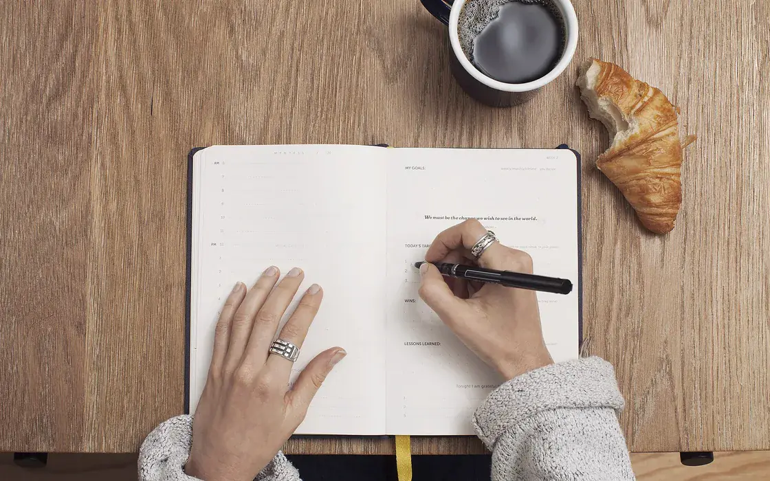 Hands writing in an open notebook on a wooden table, beside a cup of coffee.