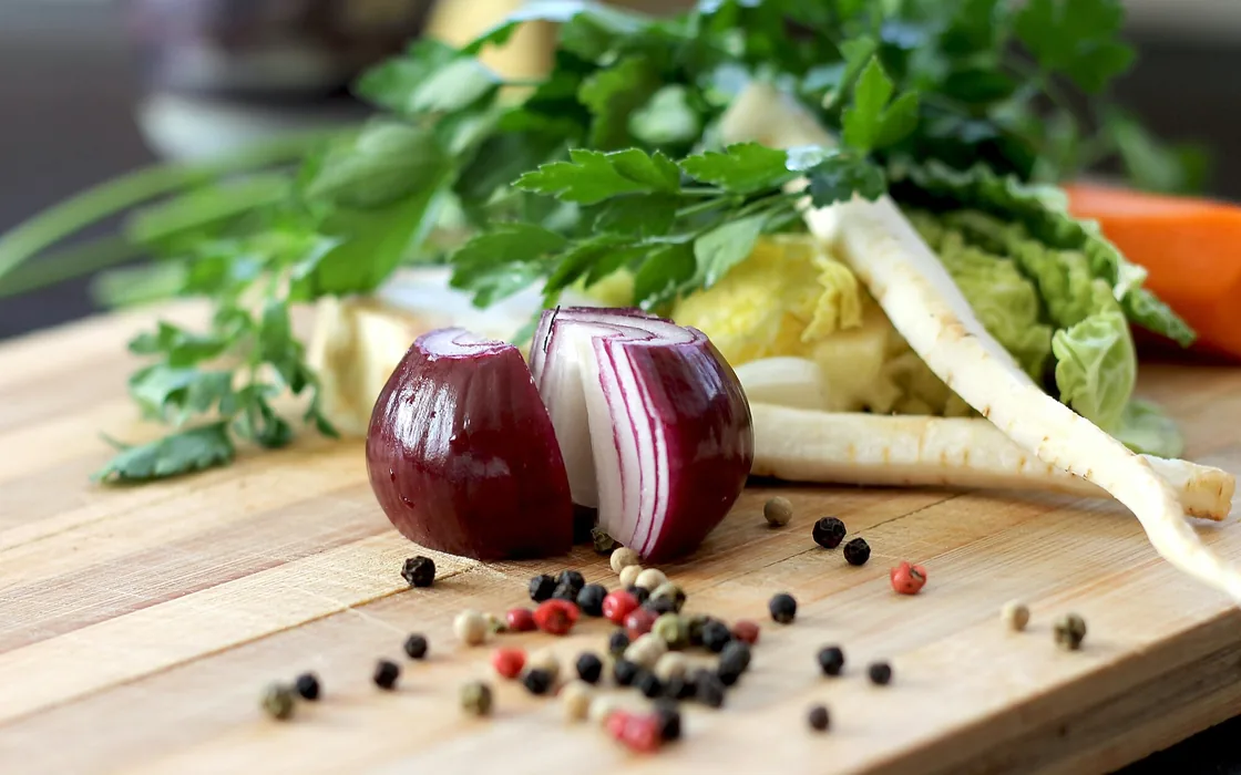 A halved red onion, herbs, parsnip and peppercorns on a wooden chopping board.