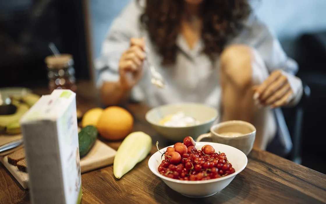 A woman at a breakfast table with yoghurt, a bowl of berries, fruit and coffee.