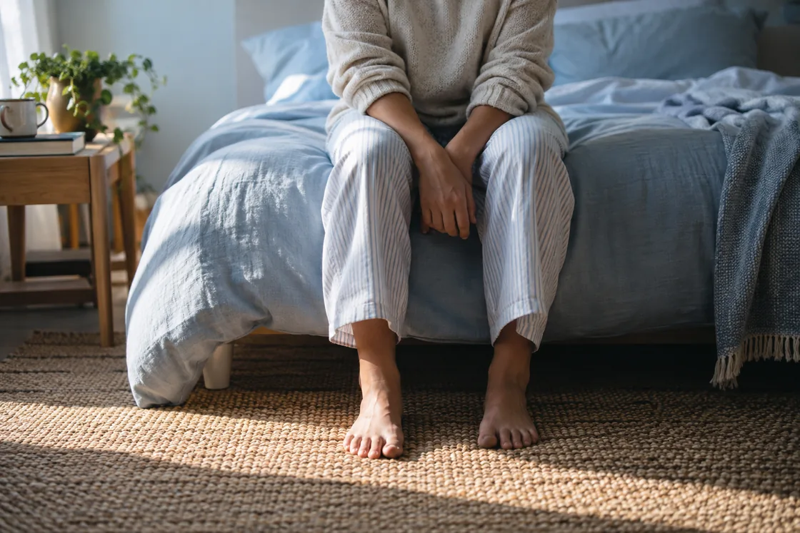 An adult sitting on the edge of a bed in morning light.